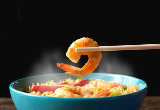 Hand using chopsticks pickup shrimp with instant noodles with smokes isolated on black background.