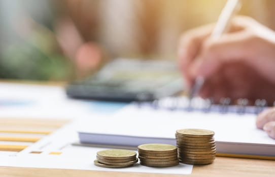 Businessman working and writing on notebook with stack of coins for financial and accounting concept.