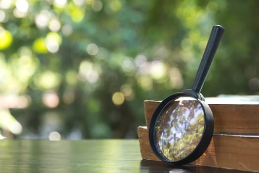 Close up of magnifying glass with old books on the wooden table and nature background.