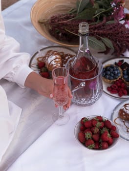 Beautiful picnic with rose wine, strawberries and berries. Female hand holding crystal glass with rose wine. Croissants, raspberries and chocolate. Bottle of wine in ice bucket