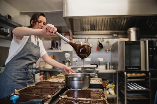 Female baker standing in a kitchen at work. She has a pan of melted chocolate and is using a kitchen utensil pour the chocolate.