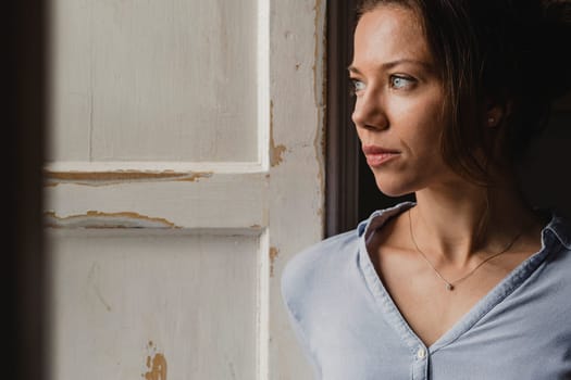 Mid adult woman standning at a wooden framed window looking out.