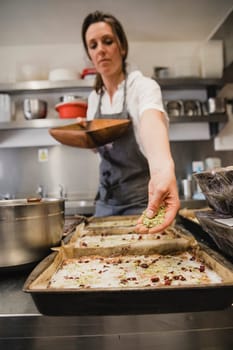 Female baker standing with her arm outstreched with crushed pistachio nuts sprinkling them onto the top of a cake.