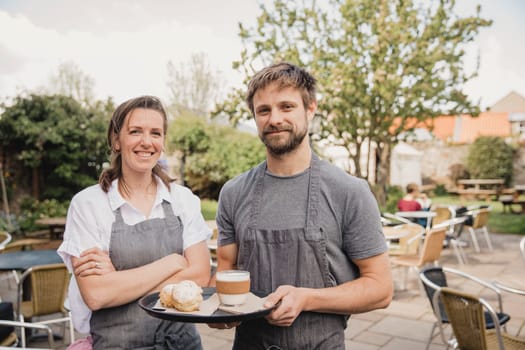 Portrait of two proud coffee shop owners standing infront of the outside seating area at their small cafe.