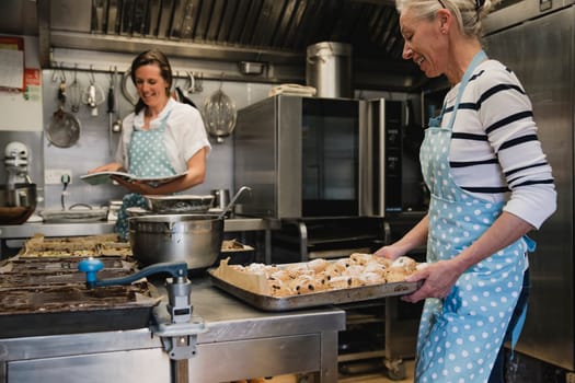 Mature female baker carrying a tray of fruit scones towards  the worktop in the kitchen.