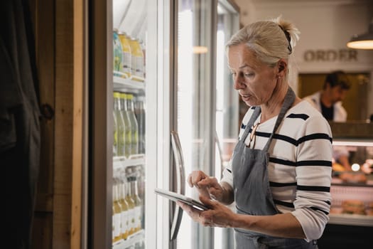 Mature coffee shop employee standing next to the fridges in the coffee shop performing a stock take.
