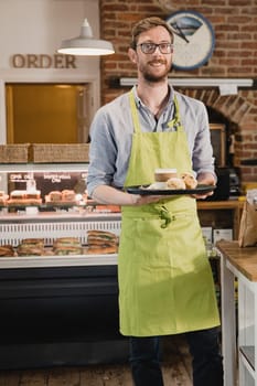 Portrait of a mid adult waiter in a coffee shop. he is looking at the camera and smiling while holding a tray with a coffee and scone on it.