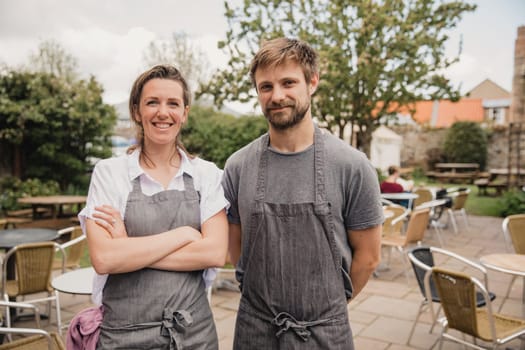 Portrait of two proud coffee shop owners standing infront of the outside seating area at their small cafe.