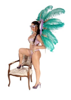 A young gorgeous woman wearing a colorful carnival outfit with big.feather, on leg on antique armchair, isolated for white background.