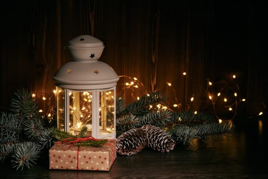 Christmas still life with fir tree branches, gift box, decorations and lamp on a dark wooden background with LED light garland, Copy space. Horisontal view. For greeting card.