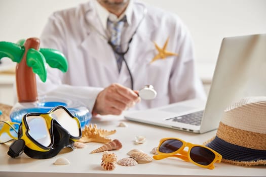 Medical and healthcare insurance for travel and summer holidays with a doctor seated at a laptop and goggles, sunglasses, sunhat and seashells in the foreground