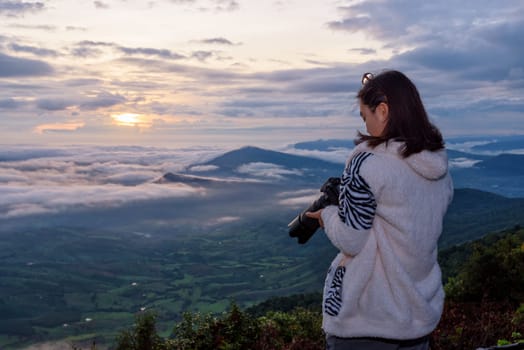 Woman tourist are looking at the camera after take a photo on nature landscape of the sun fog and mountains during the sunrise background at Phu Ruea National Park, Loei province, Thailand
