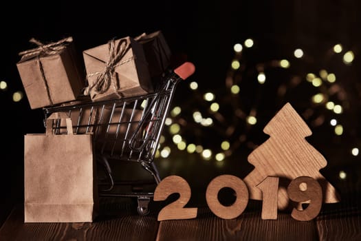 Wooden fir tree, gift boxes, shopping trolley and text 2019 from wooden figure on dark wooden background with LED light garland. Horizontal view. New Year and Christmas.