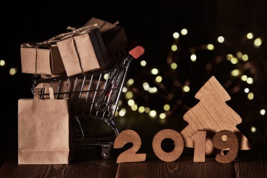 Wooden fir tree, gift boxes, shopping trolley and text 2019 from wooden figure on dark wooden background with LED light garland. Horizontal view. New Year and Christmas.