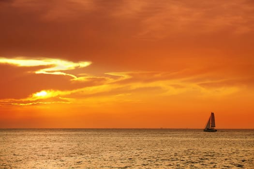 Orange color seascape image with shiny sea and sailboat over cloudy sky and sun during sunset in Cozumel, Mexico