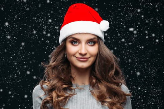 Portrait of attractive young brunette woman with long wavy hair wearing festive red christmas hat and smiling at camera over snowfall a new year night.