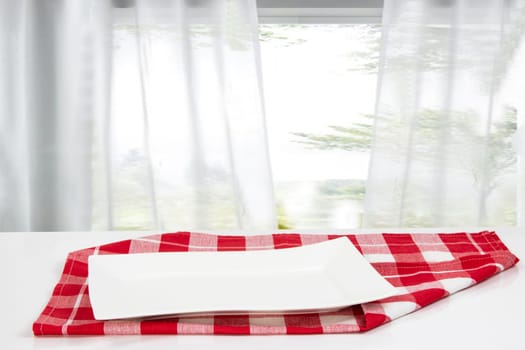 Empty plate background. Empty white plate on table with red napkin or tablecloth over blurred curtain window natural light green background. Template for your food and product display montage. Concept food. Copy space.