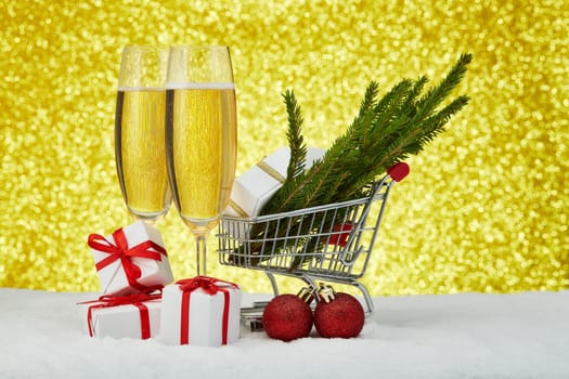 Still life with shopping trolley, gift boxes and branches of fir tree, Christmas decoration standing on snowy surface with festive gold background. Christmas and New Year sale.