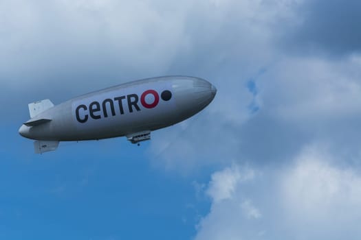 Heiligenhaus, Nrw, Deutschland - Juni 05, 2017:.Luftschiff, Zeppelin vor blauen Himmel mit dunklen Wolken und Werbebanner mit Aufschrift Centro O...HEILIGENHAUS, NRW, DEUTSCHLAND - JUNE 05, 2017:.Airship, zeppelin against blue sky with dark clouds and banners with label Centro O.