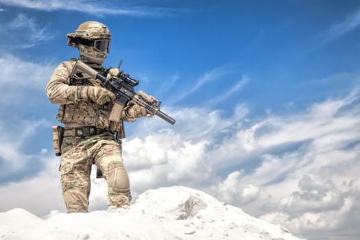 Man in military camouflage uniform and mask, equipped tactical ammunition, standing on sand dune with service rifle replica in hands, cloudy sky on background. Airsoft player taking part in war games