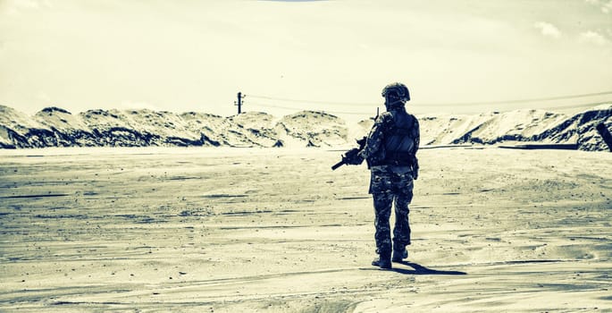 Special operations forces soldier, private military company mercenary or security armed with service rifle patrolling and observing protected territory in sandy desert area, back view, desaturated