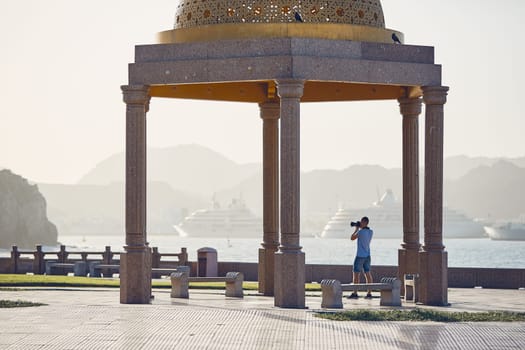 Young tourist photographing waterfront against harbor with modern passenger ships. Mutrah Corniche in Muscat, Sultanate of Oman.
