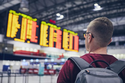 Rear view of young man checking flight schedule on the arrival departure board at airport.