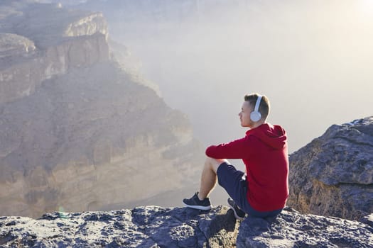 Relaxation in mountains. Young man with headphones sitting on the edge of cliff and listening music.