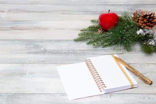 Christmas decoration background. Composition from sparkling christmas tree branches with a red apple and fir cones and a blank notebook with pen on a rustic wooden background. Top view copy space.