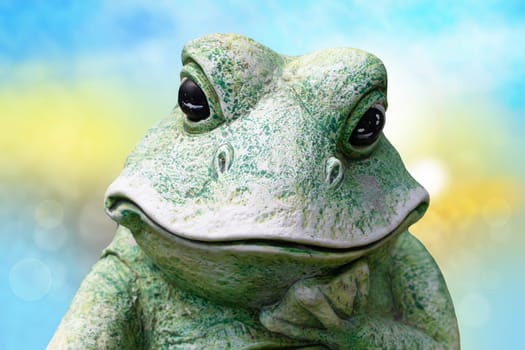 Close-up of frog head. A decorative old weathered ceramic frog over abstract blue yellow pastel background. Ceramic frog decoration.