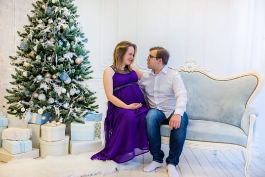 Pregnant woman in ultra violet dress with husband sitting on the sofa near christmas tree