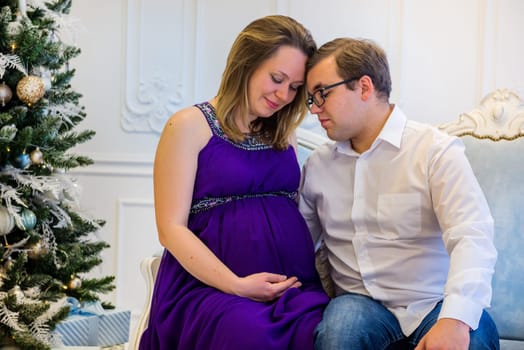 Pregnant woman in ultra violet dress with husband sitting on the sofa near christmas tree