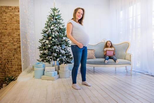 Beautiful pregnant woman standing near christmas tree with daughter sitting on the sofa