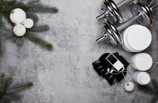 Sports nutrition (supplements), dumbbells, fir tree branches and christmas ornament on a gray background. Top view with copy space. New Year and Christmas. Fitness. Healthy lifestyle.
