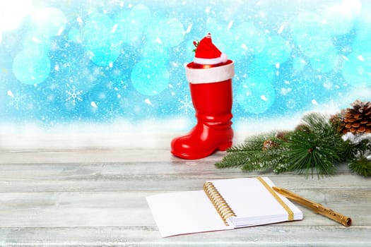 Christmas decoration background. Composition from christmas tree branches with a red Santa boots and a blank notebook with pen on a rustic wooden table over blue abstract snowy bokeh background. Space.