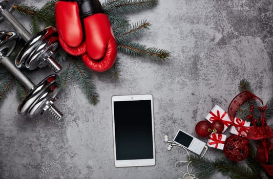 Dumbbells, boxing gloves, computer tablet, fir tree branches and Christmas decorations on a gray background.Top view with copy space. New Year and Christmas. Fitness, sport and healthy lifestyle concept.