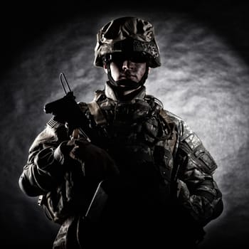 Low key studio portrait of private security service contractor, army infantry rifleman, US marine raider in helmet, sunglasses, camouflage uniform posing with weapon on black background with backlight