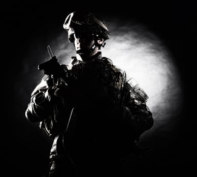 Low key studio portrait of private security service contractor, army infantry rifleman, US marine raider in helmet, sunglasses, camouflage uniform posing with weapon on black background with backlight