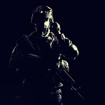 US marine raider in combat uniform with hidden face, armed with assault carbine low key, high contract studio shot on black background. Equipped army soldier standing in darkness with weapon in hands