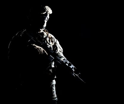 US marine raider in combat uniform with hidden face, armed with assault carbine low key, high contract studio shot on black background. Equipped army soldier standing in darkness with weapon in hands