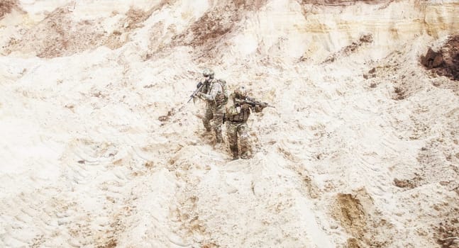 Two infantry soldiers in combat camo uniform, with tactical ammunition, carefully walking and aiming with assault rifles in unknown desert area. Military reconnaissance, scouting on enemy territory