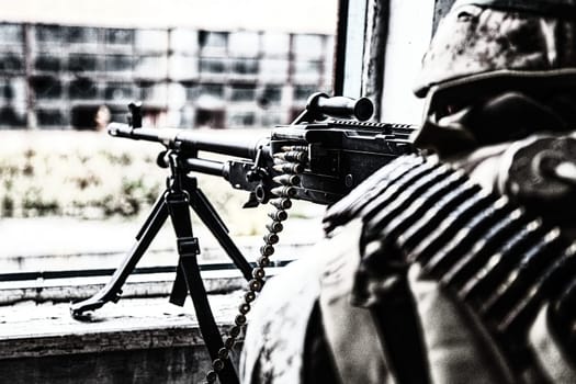 Commando machine gunner, U.S. Marine Corps infantryman, military squad automatic weapon operator in helmet and ammo belt around body, firing through window in ruined building, over shoulder back view