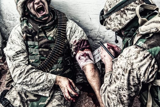 Marine wounded in shoulder, suffering of pain and screaming while receiving medical aid from comrade. Military medic apply pressure bandage to casualty, binding gunshot wound, trying stop bleeding