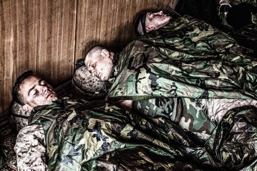 Tired U.S. marines resting at temporary base or camp, lying on floor in uniform and tactical ammunition, covered with poncho liners and sleeping bags, sleeping well after hard raid, exhausting mission