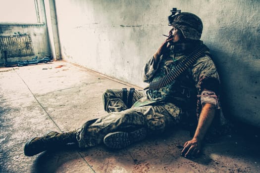 Soldier in combat uniform and ammo belt over chest, sitting on floor and smoking after being shoot in shoulder and put bandage on wounded hand, dropped weapon lying nearby with blood stains around