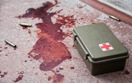 First aid kit metal box lying on floor with empty bullet shells and fresh stains of human blood around. Emergency medical care for gunshot wounds, bleeding stop, tactical combat casualty care concept