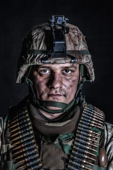Shoulder portrait of experienced army soldier, military conflict veteran, skilled marine fighter in ragged camouflage uniform, advanced helmet and ammo belts on chest, studio shot on black background