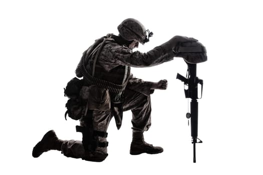 Army soldier in sorrow for fallen comrade, standing on knee, leaning on rifle with helmet and two dog tags on chain, studio shoot isolated on white low key silhouette. Military funeral honors, grief for killed in action