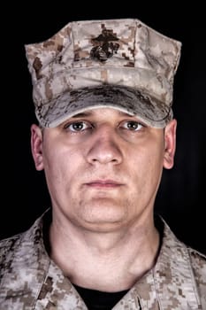 Close up studio portrait of U.S. marines infantry soldier in camouflage combat uniform and patrol cap with United States Marine Corps emblem on it, looking at camera, isolated on black background