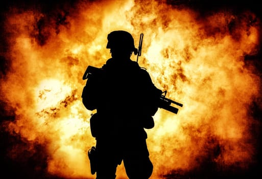 Silhouette of modern infantry soldier, elite army fighter in tactical ammunition and helmet, standing with assault service rifle in hands on background of fiery explode. Burning fire of war conflict